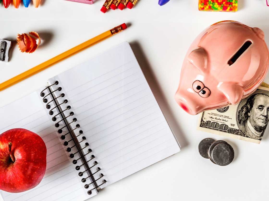 A notebook with apple and pencil next to a piggy bank, cash and coins.